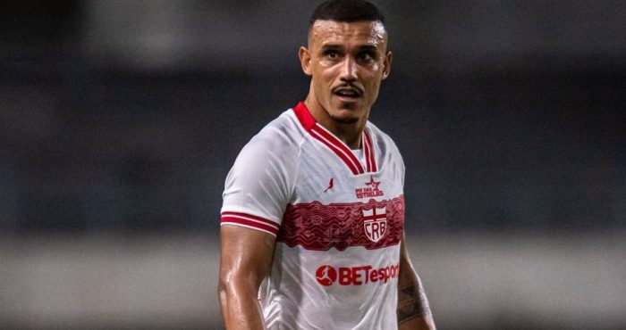 "Fotografia colorida do jogador William Pottker vestindo o uniforme branco do CRB. Ele é um homem pardo, com cabelo curto cortado em degradê e um bigode fino. O atleta aparece suado, com expressão focada, olhando para o lado durante uma partida noturna. A camisa possui detalhes em vermelho na gola e nas mangas, com o escudo do clube e o patrocínio 'BETesporte' em destaque."