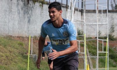 Foto de meio corpo do zagueiro Edson Rogério durante um treino ao ar livre. Ele veste uma camisa de treino azul clara com detalhes em azul marinho e o patrocínio "Estrela Bet". O jogador está em movimento de corrida, focado, com um campo gramado e uma estrutura metálica branca ao fundo.