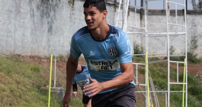 Foto de meio corpo do zagueiro Edson Rogério durante um treino ao ar livre. Ele veste uma camisa de treino azul clara com detalhes em azul marinho e o patrocínio "Estrela Bet". O jogador está em movimento de corrida, focado, com um campo gramado e uma estrutura metálica branca ao fundo.
