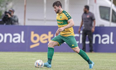 "Zagueiro Gabriel, do Cuiabá EC, conduz a bola durante uma partida. Ele veste o uniforme amarelo e verde do Dourado, com o patrocínio 'Betnacional' visível ao fundo. O jogador demonstra concentração e postura firme no campo, enquanto um membro da equipe técnica e uma câmera aparecem ao fundo, registrando o momento."