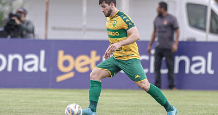 "Zagueiro Gabriel, do Cuiabá EC, conduz a bola durante uma partida. Ele veste o uniforme amarelo e verde do Dourado, com o patrocínio 'Betnacional' visível ao fundo. O jogador demonstra concentração e postura firme no campo, enquanto um membro da equipe técnica e uma câmera aparecem ao fundo, registrando o momento."